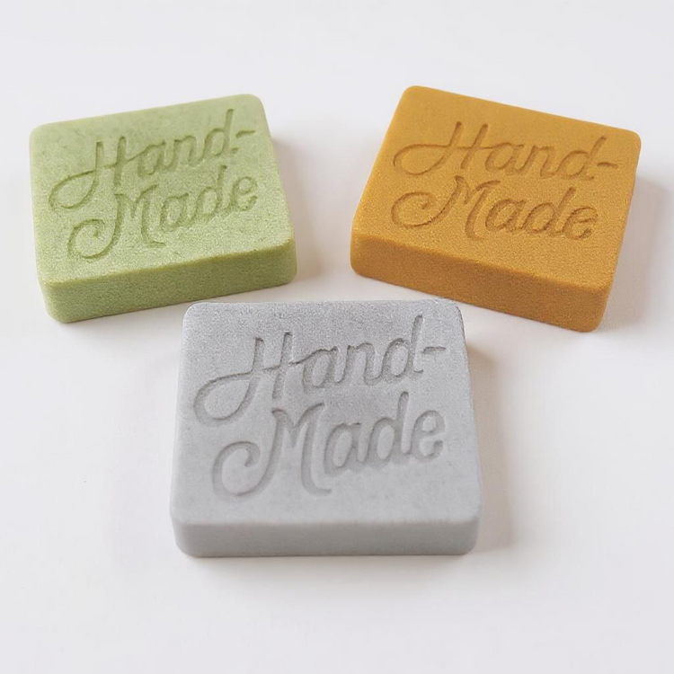 To soap bars one tinted green one tinted Gray on a white background both with paper Brown wrappings with labels