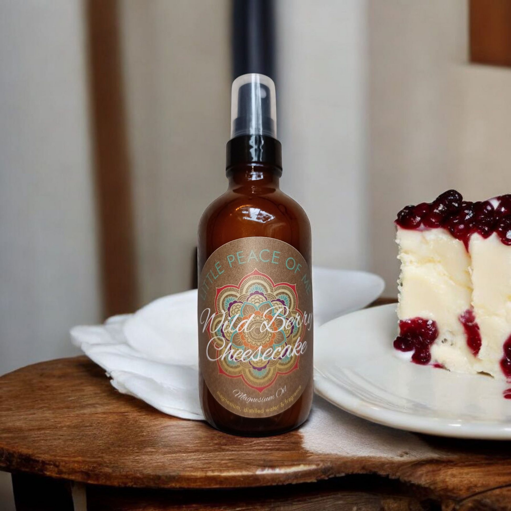 4 oz glass spray bottle that reads the logo a Little piece of Me wild berry cheesecake magnesium oil sitting on a wooden table ledge with a napkin behind it with a plate sitting next to it with a large piece of cheesecake with berry toppings with a backdrop a part of the kitchen with white and dark walls