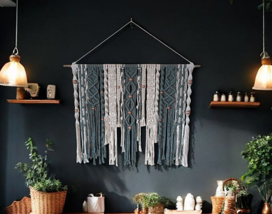 A dark tealish green and white macrame wall hanging intrically nodding strings hanging down off of a wooden rod hung on a wall backdrop has a couple of shelves with some decorations and a shelf below the wall hanging with a plant and some other decorations on a dark background wall also two lights coming down from the ceiling on both sides