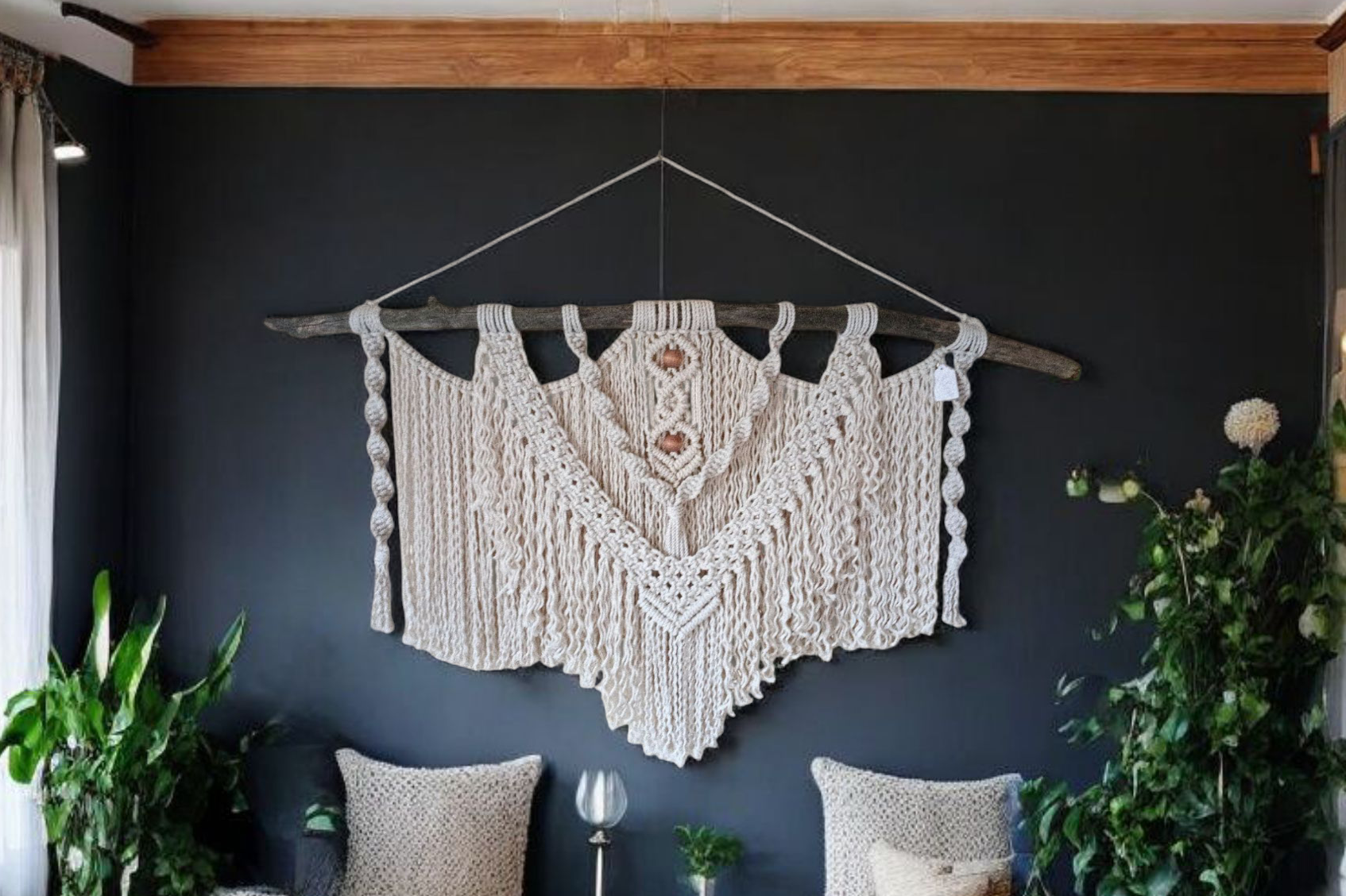 A large macrame wall hanging with white intricate nodding with strings hanging down giving it Bohemian look hanging on a large piece of driftwood hanging on a wall that is dark with wood trim running along the ceiling a few pillows underneath it with some decorations and candles plants running up the wall on both sides 48 in Long 20 in height on the macrame measurements