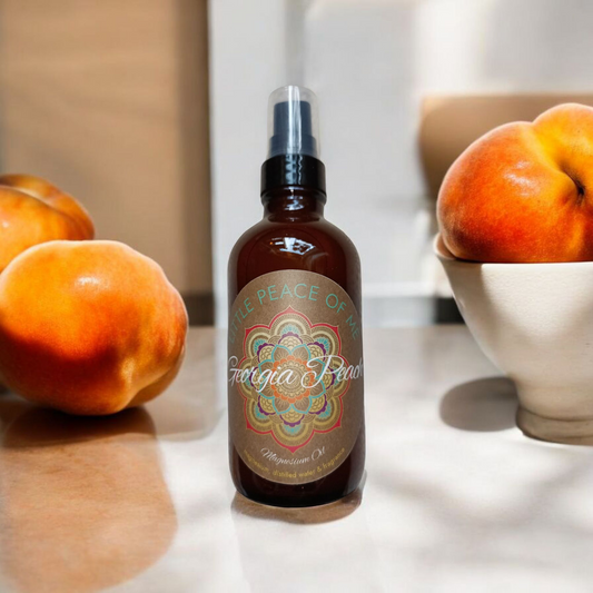 4 oz spray bottle that reads little piece of me Georgia peach magnesium oil spray sitting in a countertop with lighting shining through a window onto two large peaches on one side and a bowl that's white with some peaches in it displayed next to the bottle a wall runs the up close next by to it with tan paint
