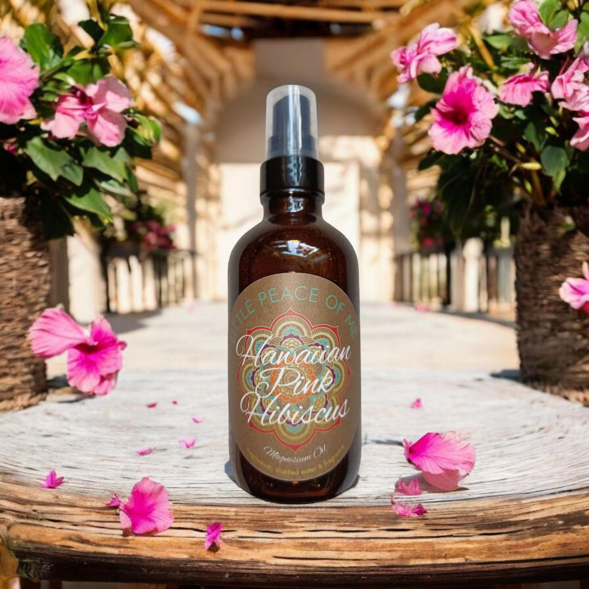 4 oz Boston Brown spray bottle with a tan label that says little piece of knees Hawaiian pink hibiscus sitting on a ledge a brown slate that runs along the backdrop of a large room on both sides of the bottle are hibiscus plants with a few hibiscus flowers laying next to a bottle