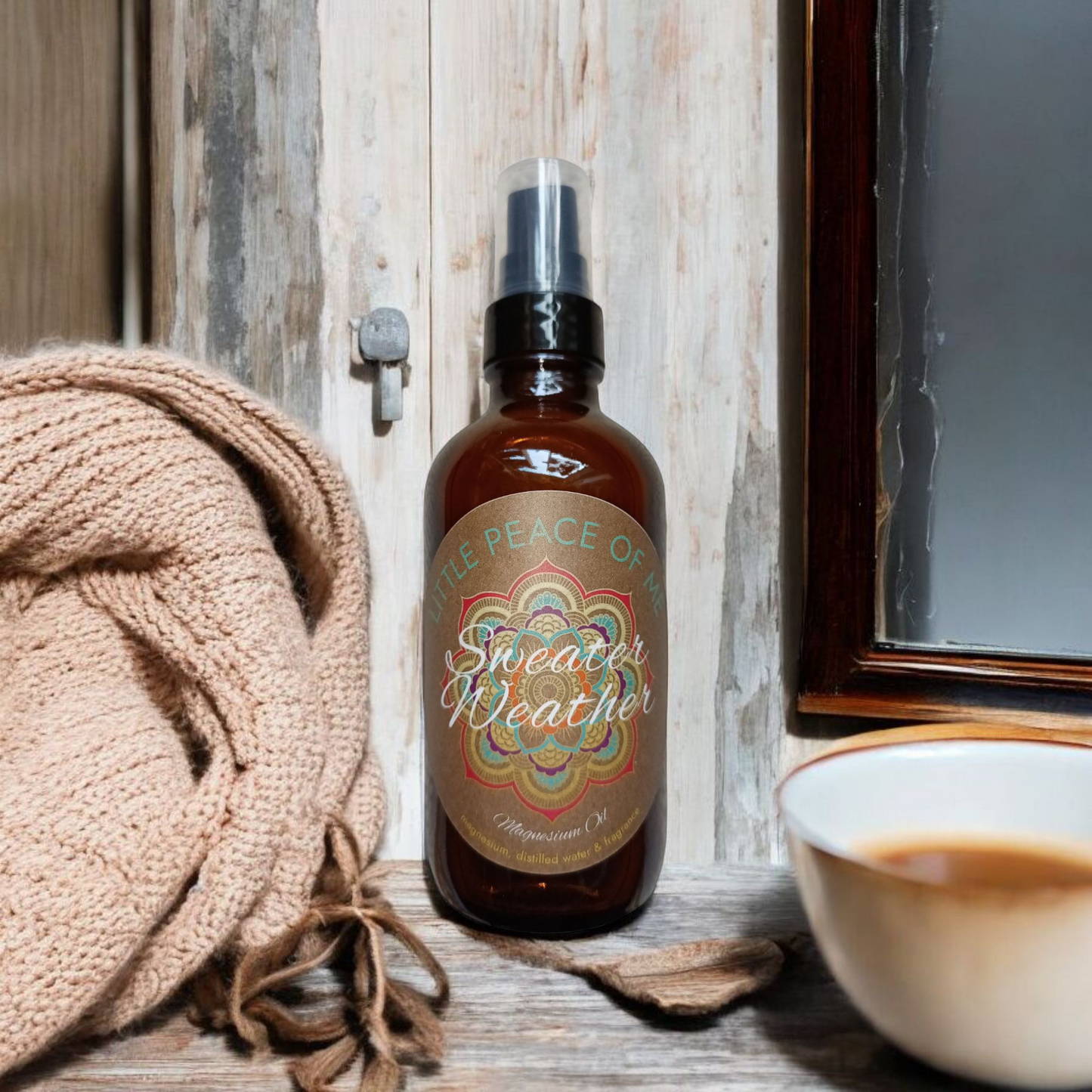 Centered in the center is 4 oz Boston spray bottle darken color with the label that's tan that says little piece of knees sweater weather magnesium oil sitting back a little from a cup of coffee and off to the other side is a sweater wrapped up up against a wall that has a window and white wood up the wall