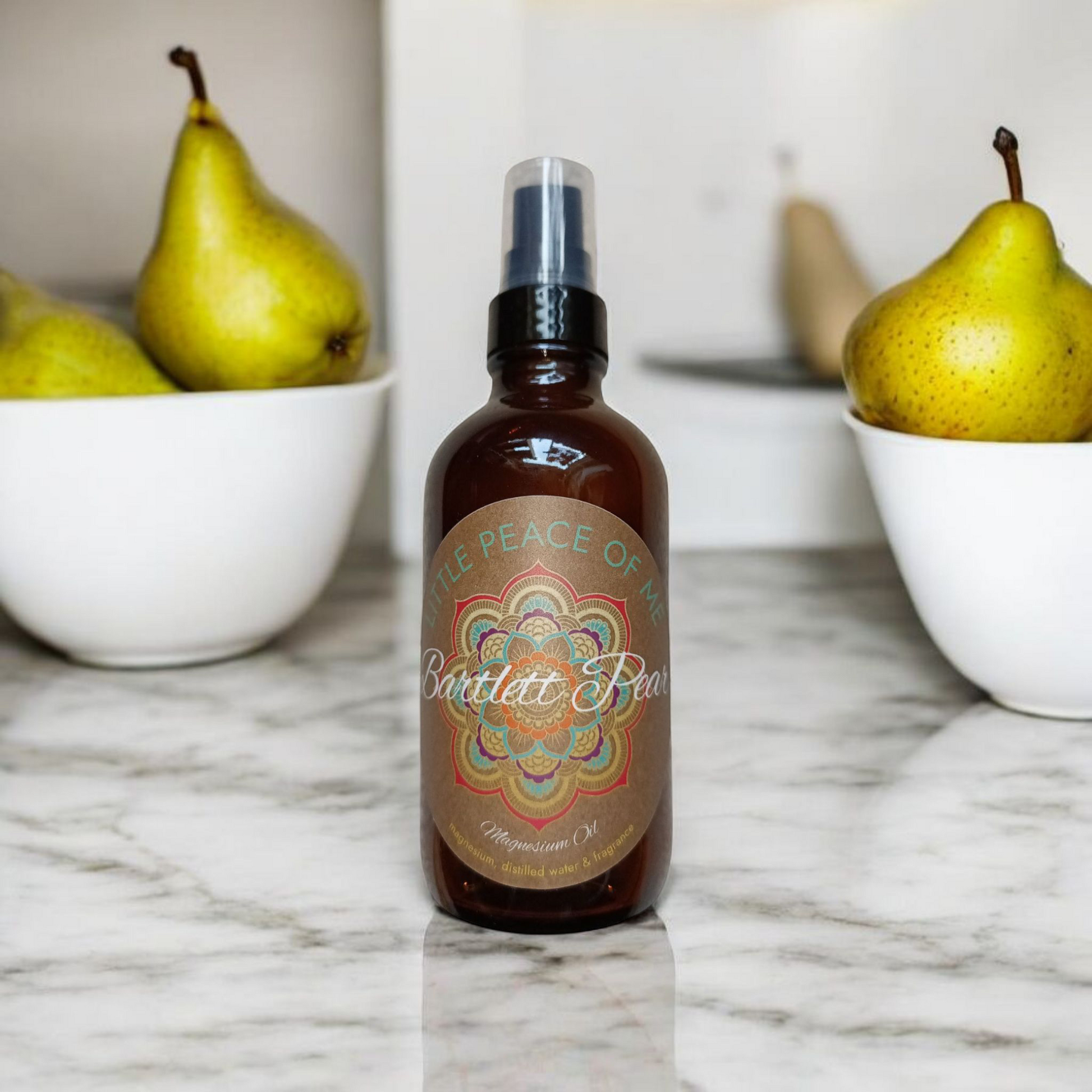 4 oz Brown spray bottle with the logo that reads Little piece of Me the scent is Bartlett Pear magnesium oil sits on a marble countertop setback behind it are two bowls full of pears on a whitewalled backdrop in a kitchen