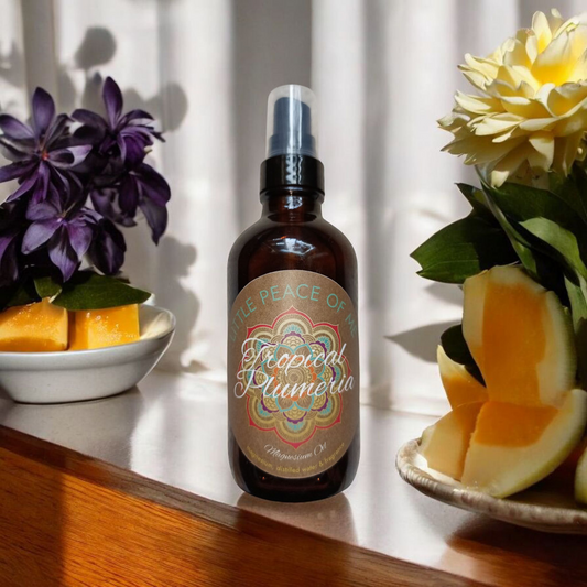 4 oz dark Boston spray bottle with a tan label that says Little piece of Me tropical Plumeria magnesium oil sitting on a ledge with the sun shining through onto it displayed in front side and back side are some flowers and fruit displayed on a white wall with sunlight shining on it