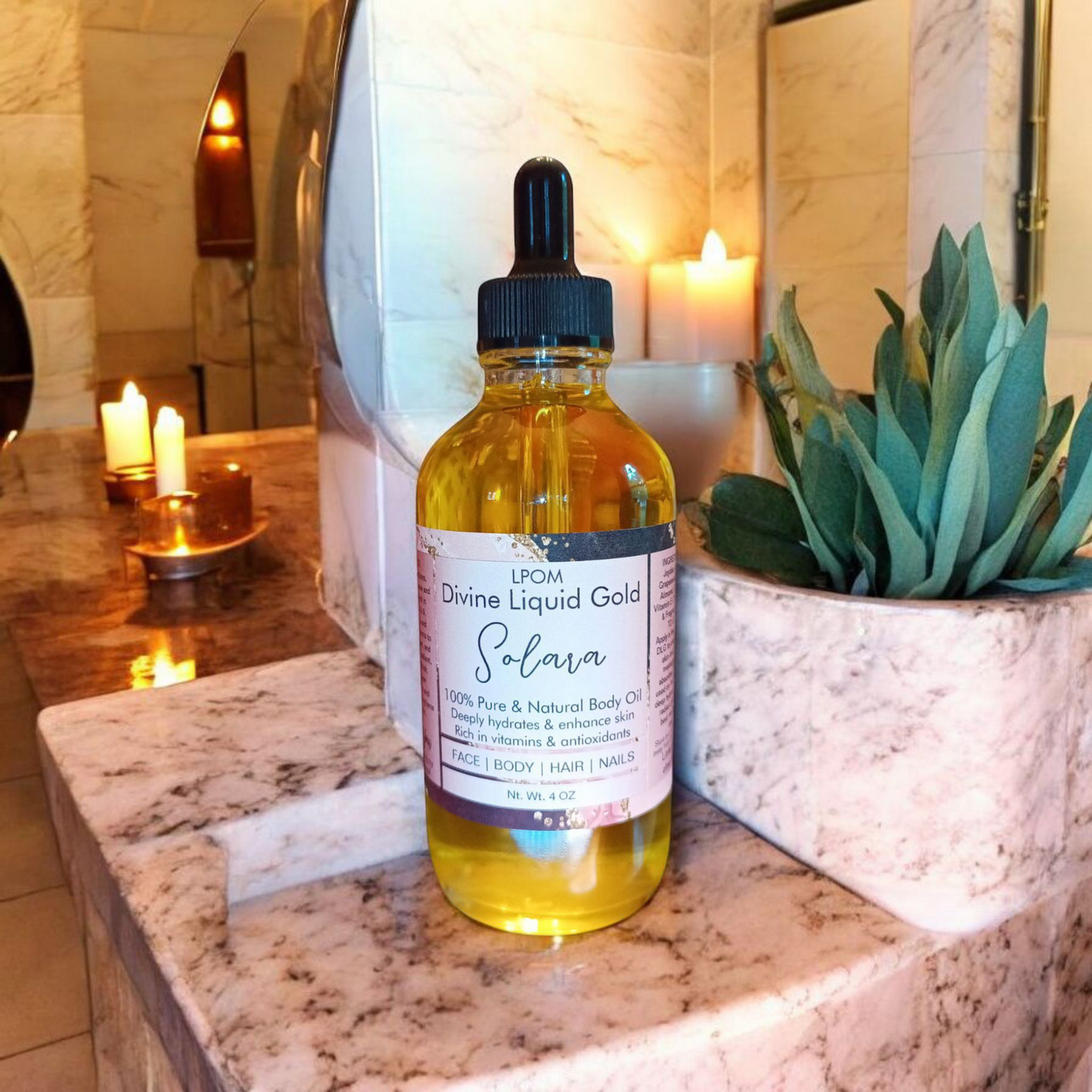 Forearms dropper bottle clear glass with a label that says Divine Liquid Gold body oil scent name Solara sitting on a tan marble counter alongside of some candles and a small plant background of a small bathroom with some lighting