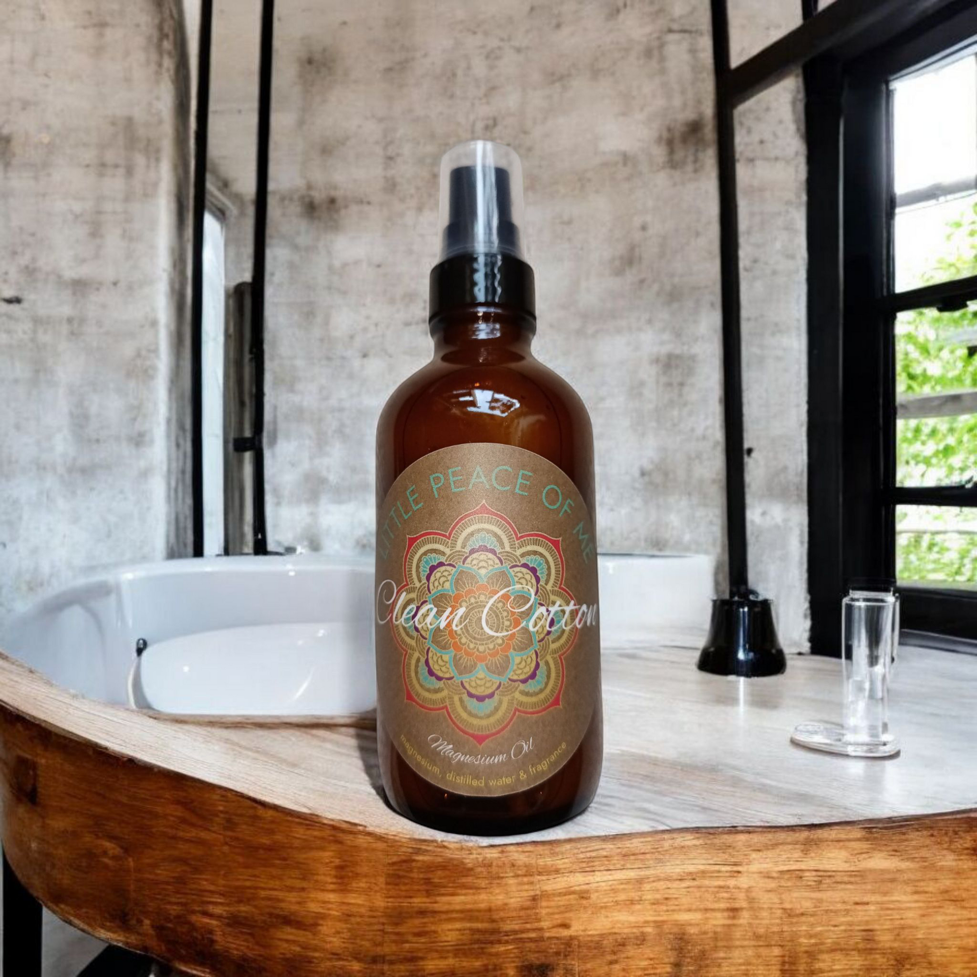 4 oz dark Boston spray bottle with tan label that reads little piece of me clean cotton magnesium oil sitting on a counter ledge that's white with wooden accents on a cement wall bathroom with industrial metal accents with a towel hanging set behind the bottle in a bathroom setting with a window off to the side