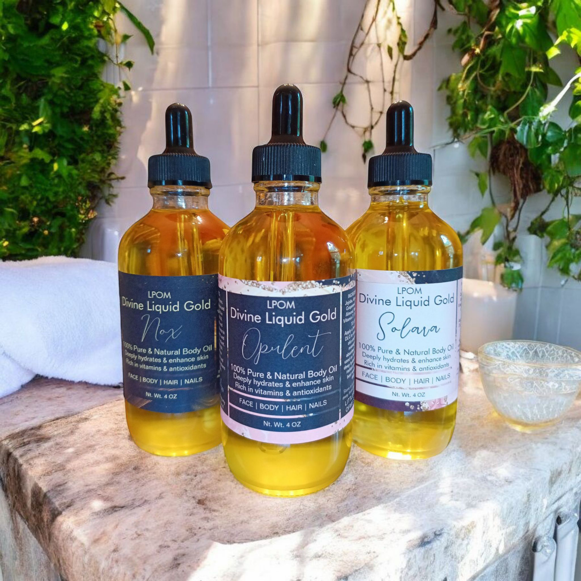 Three 4 oz bottles glass droppers with labels that read the vine Liquid Gold and their scents played on a marble backdrop towels to the side one of the bottles plants run up the back wall
