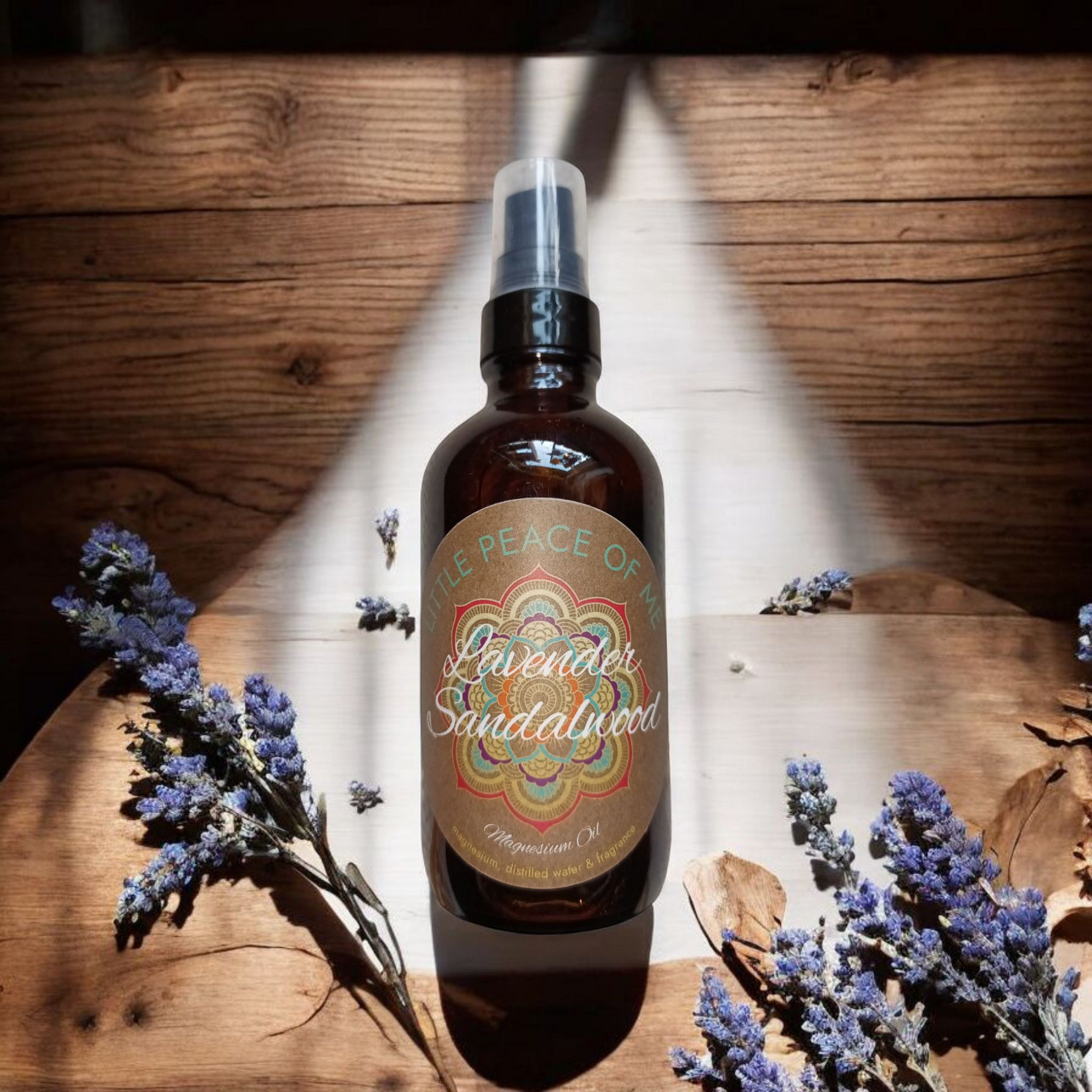 4 oz spray Boston dark bottle with a label that's tan that says little piece of me lavender sandalwood laid down on its side on a wooden table with some dried lavender laying next to it sun shining casting a light across it