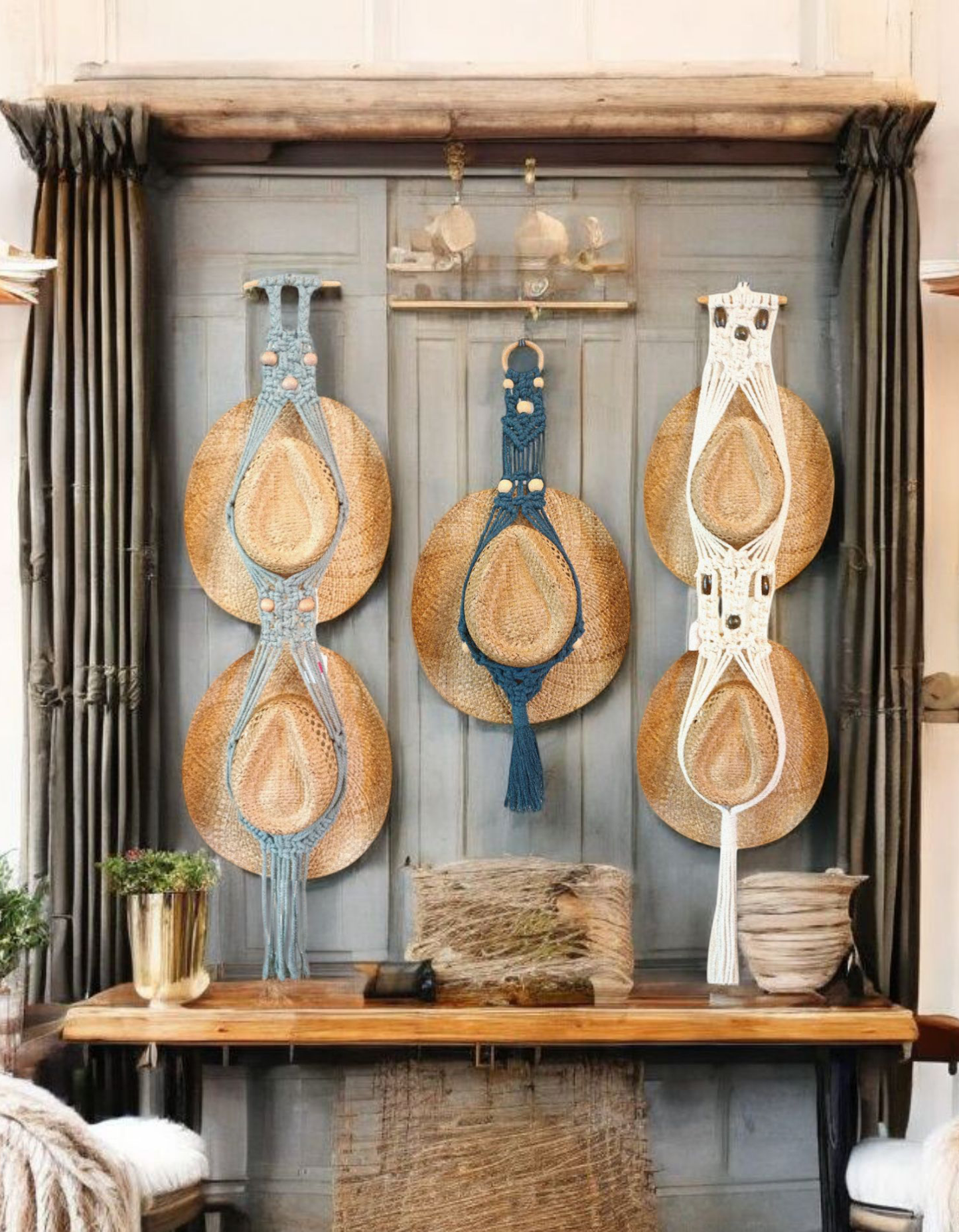 Zoomed in backdrop of a doorway with a wall with a small bench sitting at the bottom with a few baskets and other decorations all displayed around three macrame hat holders the macrames are intrically knotted to hold hats one is a light teal with some beads that are white one is black with some beads and another one is white with some dark beads sitting in a country little setting