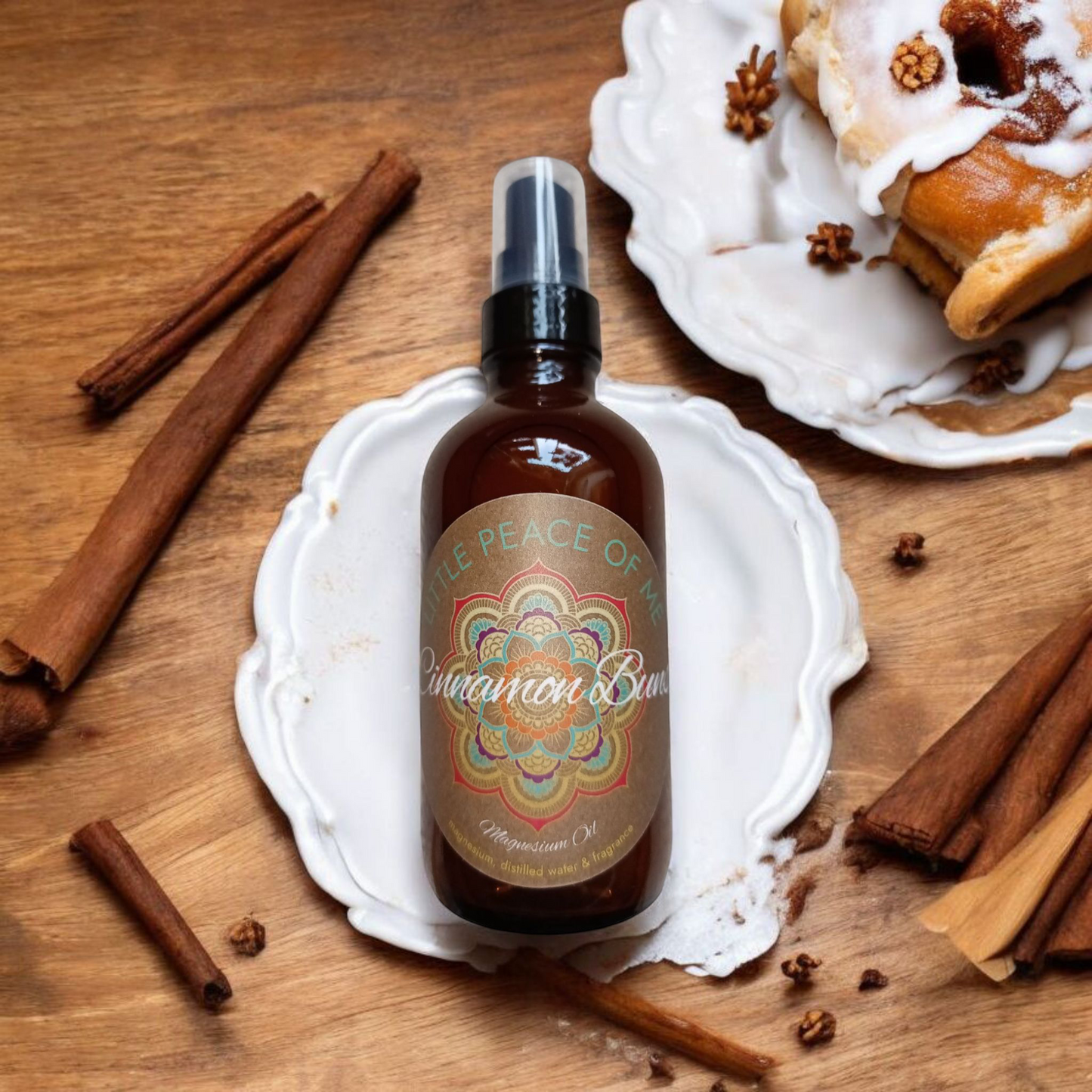 A 4 oz Boston spray bottle with a label that's tan that says little piece of me is cinnamon buns magnesium oil that's laying on a white icing displayed on a wooden table with a plate that has a cinnamon bun with icing and cinnamon sticks are displayed out around the bottle on the table